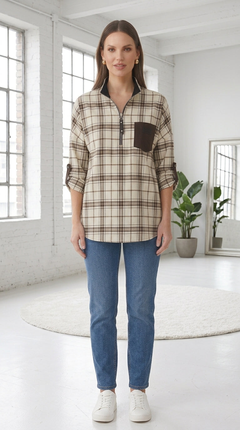 A woman modeling a beige and brown plaid button-up shirt with a half-zip collar and a chest pocket, paired with blue jeans and white sneakers in a bright, modern studio setting.