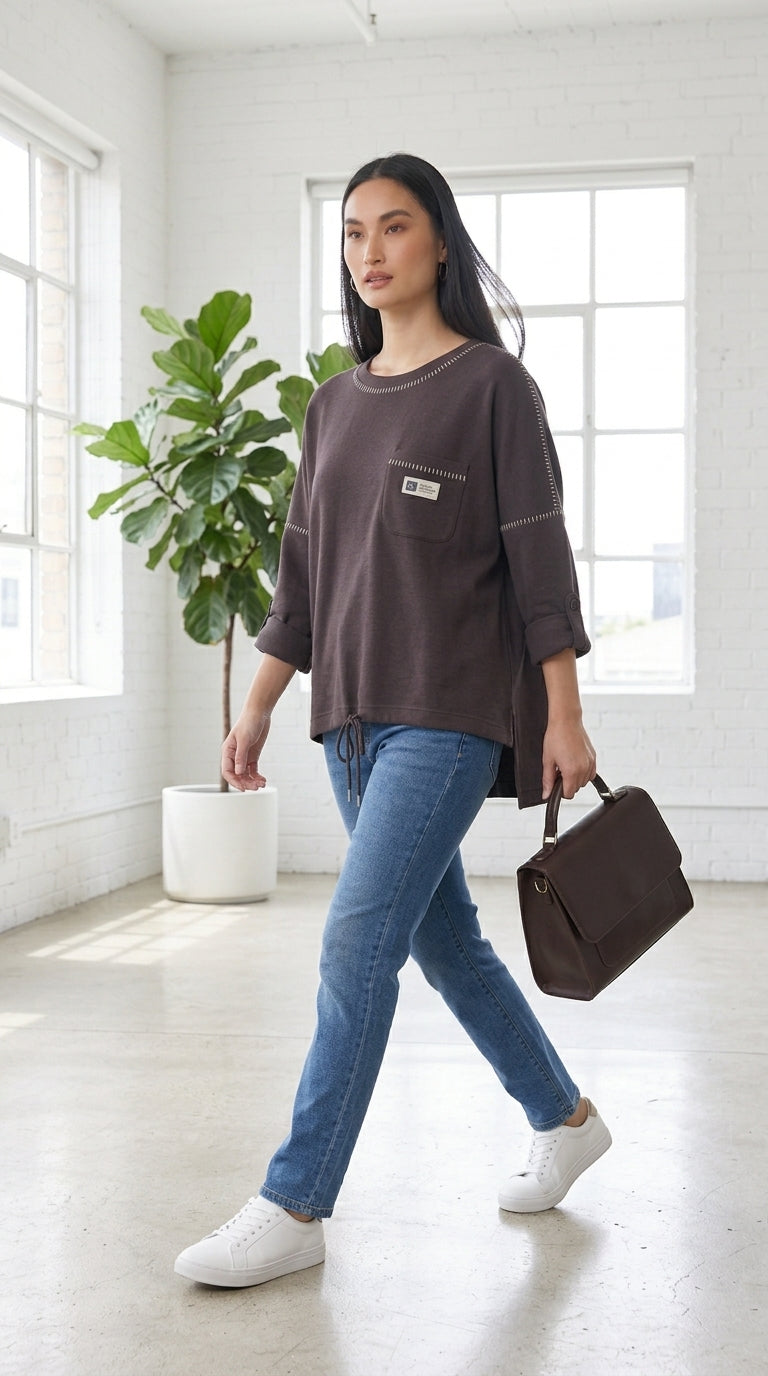 A woman walks indoors wearing a dark brown oversized long-sleeve top with a chest pocket, blue jeans, and white sneakers, while carrying a dark brown handbag in a bright, modern room with large windows and a potted plant.