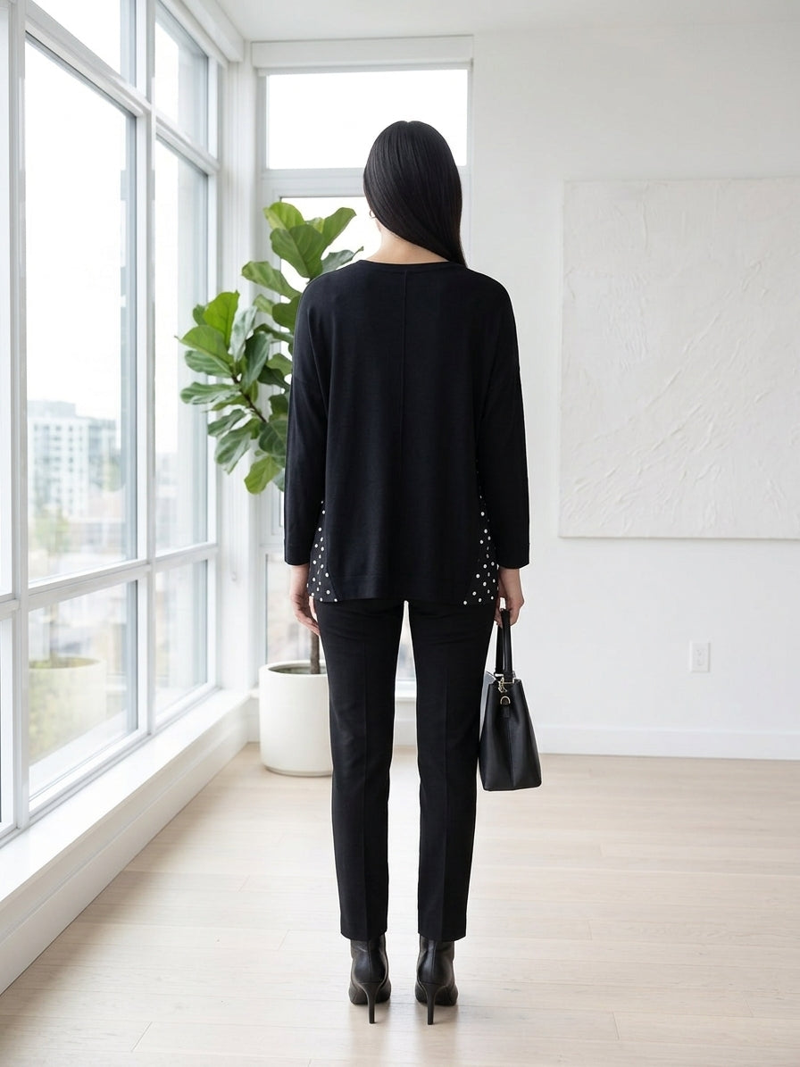 A woman stands with her back to the camera in a bright, modern room, wearing a black long-sleeve top with silver beaded details on the sides, black pants, and black ankle boots. She is holding a black handbag.