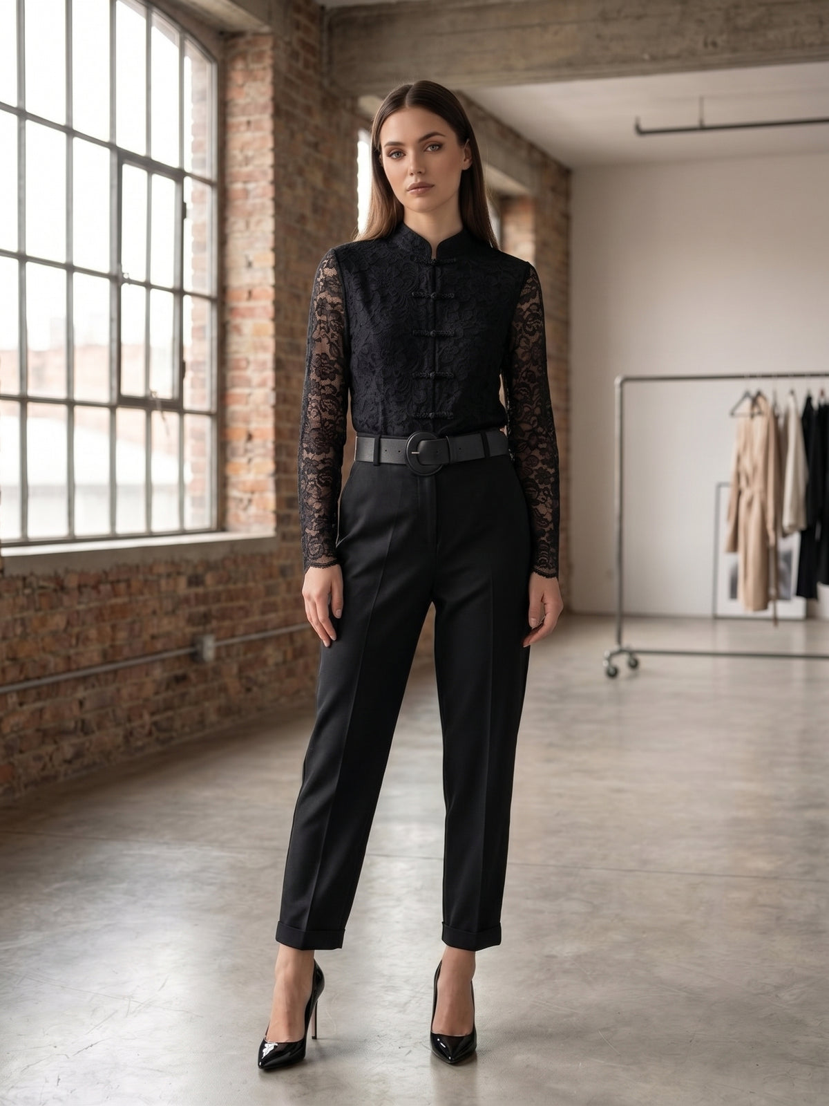 A woman stands in a loft-style room with exposed brick walls and large windows, wearing a black lace-trimmed mock-neck top with long sleeves and black trousers, paired with black high heels.