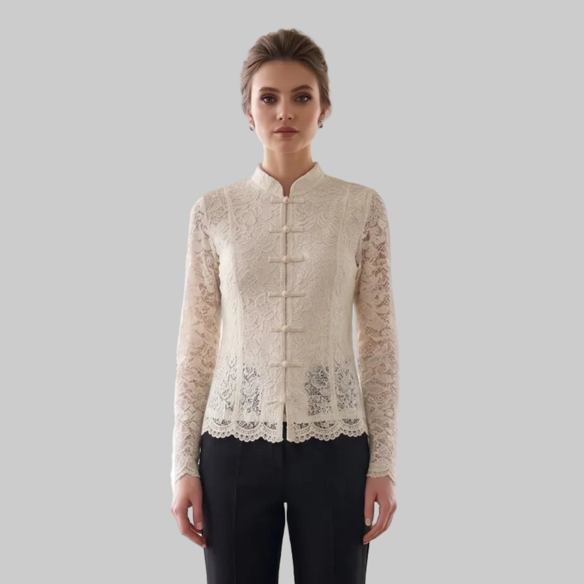 A woman stands in a bright, modern studio with white brick walls, wooden ceiling beams, and potted plants. She is wearing a cream-colored lace long-sleeve top with a mandarin collar and black trousers.
