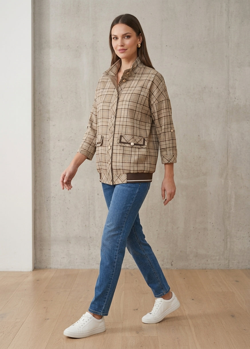 A woman modeling a beige and brown plaid button-up shirt jacket with three-quarter sleeves, paired with blue jeans and white sneakers, standing in a room with a concrete wall and wooden floor.