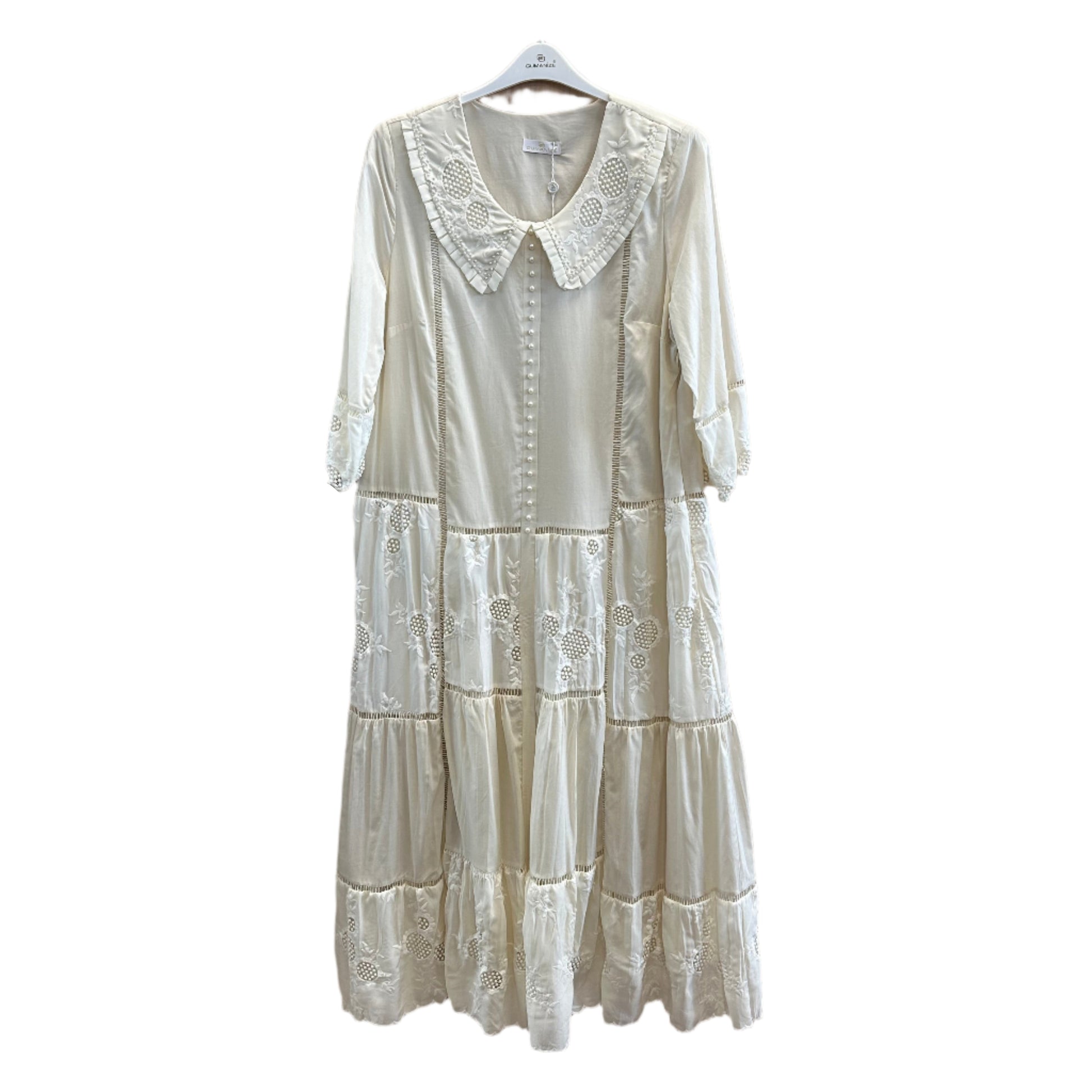 Ilga bohemiško stiliaus suknelė su nėriniuota apykakle ir perliukų sagomis, dekoruota siuvinėtais intarpais, pakabinta ant pakabos.