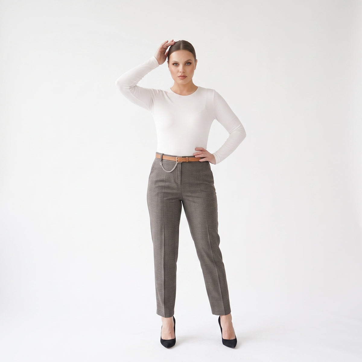 A woman modeling a white long-sleeve crewneck top and gray tailored trousers with a thin brown belt and silver chain detail, paired with black heels, against a plain white studio background.