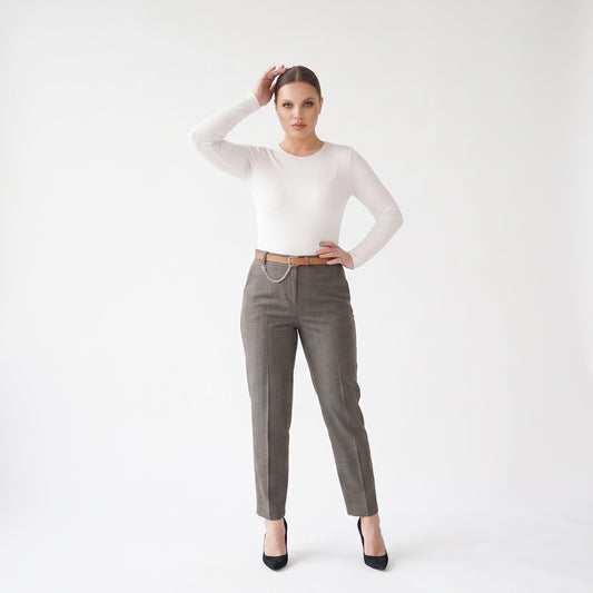A woman modeling a white long-sleeve crewneck top and gray tailored trousers with a thin brown belt and silver chain detail, paired with black heels, against a plain white studio background.