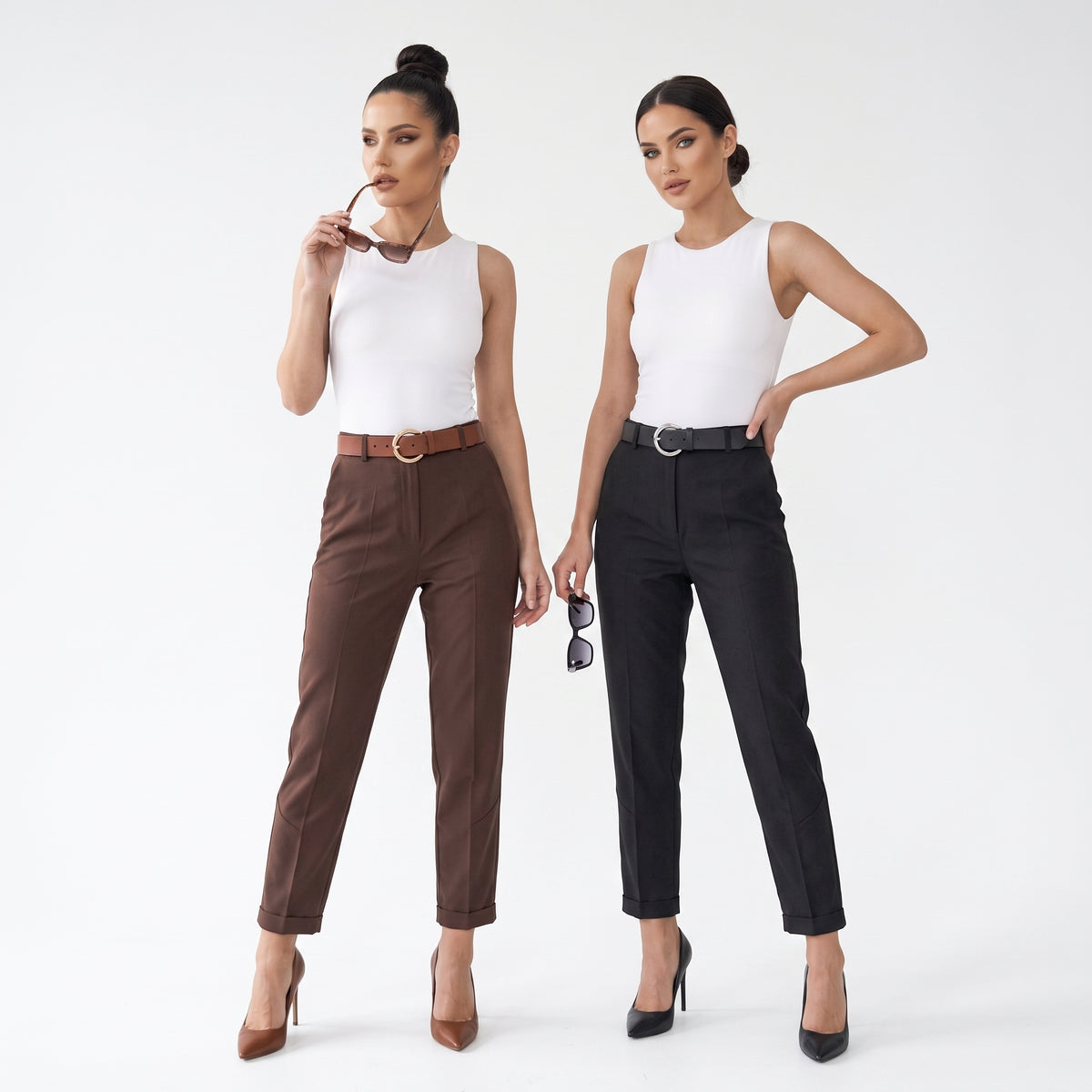 Two women modeling straight-leg trousers in a studio setting. The woman on the left wears brown trousers with a brown belt and a white sleeveless top, while the woman on the right wears black trousers with a black belt and a white sleeveless top.