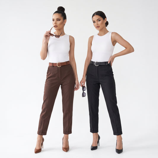 Two women modeling straight-leg trousers in a studio setting. The woman on the left wears brown trousers with a brown belt and a white sleeveless top, while the woman on the right wears black trousers with a black belt and a white sleeveless top.