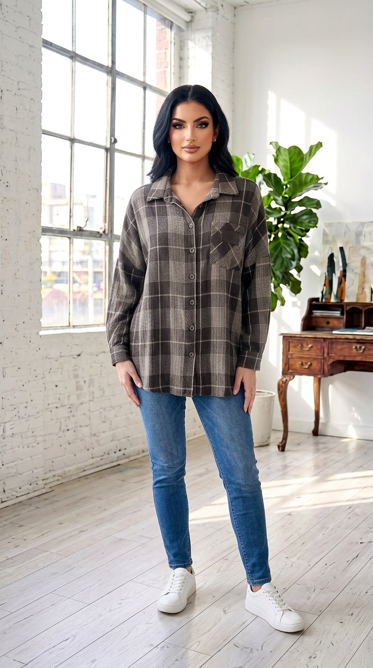 A woman modeling a brown and gray plaid button-up shirt, paired with blue skinny jeans and white sneakers, standing in a bright, modern loft room with white brick walls and a large window.