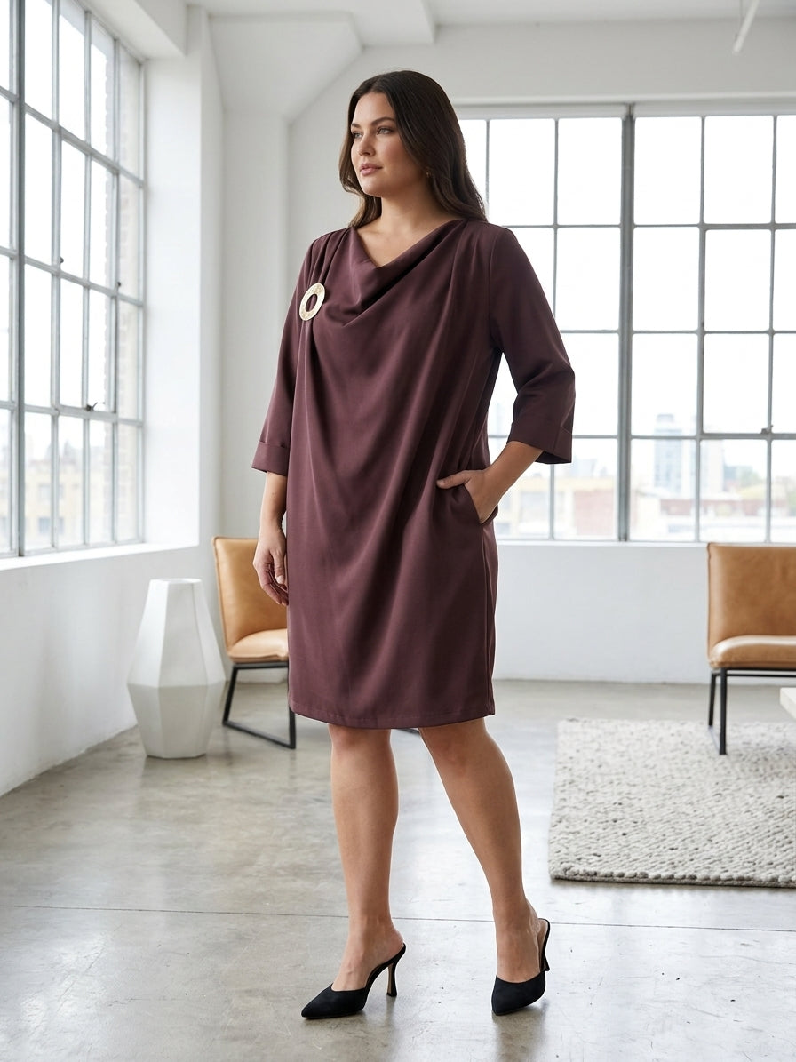 A woman models a dark plum-colored cowl-neck shift dress with three-quarter sleeves and side pockets, styled with black pointed-toe heels in a bright, modern loft setting with large windows.