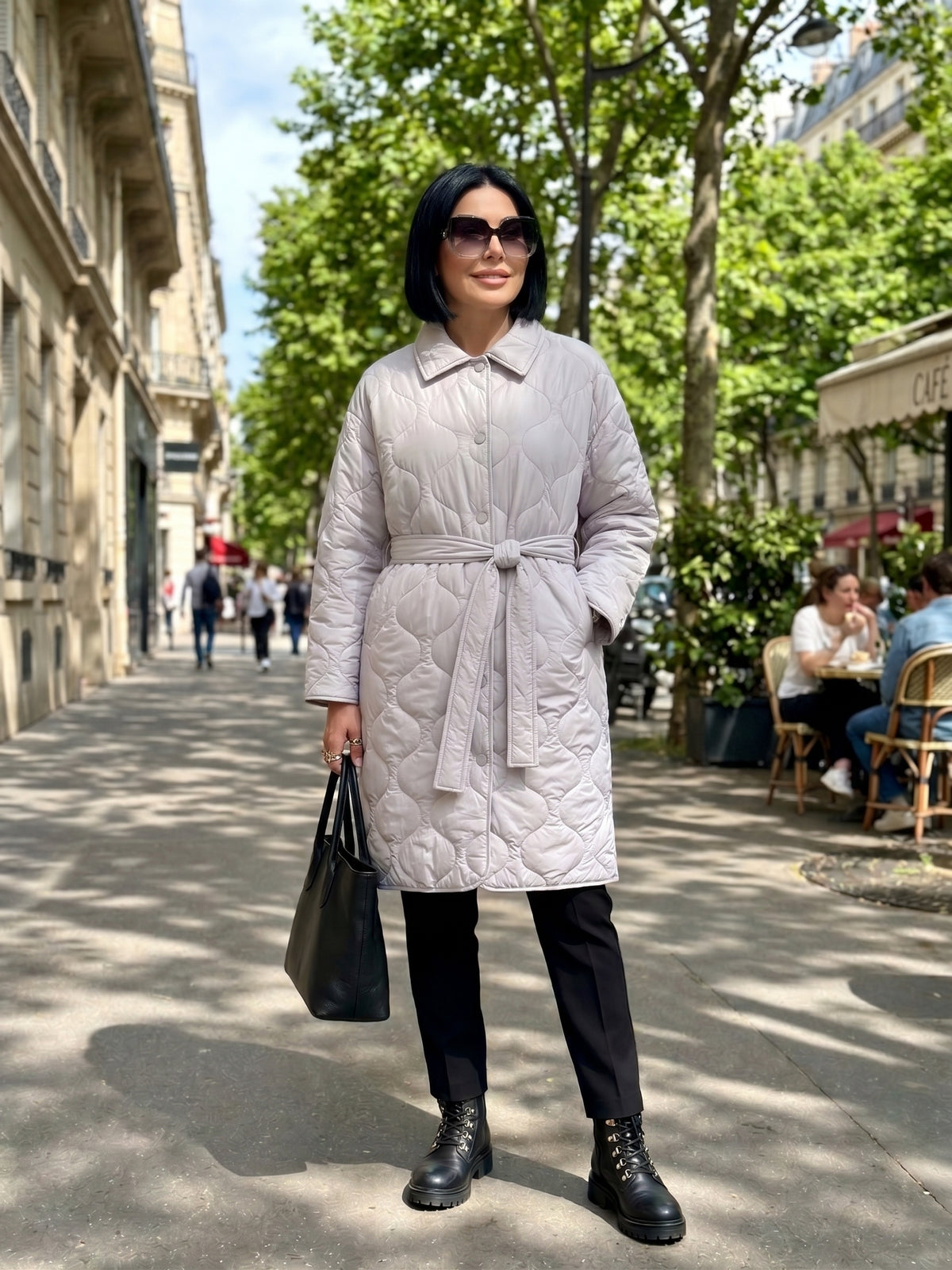 A woman stands on a sunny city sidewalk wearing a light gray quilted belted coat, black pants, black boots, and holding a black tote bag. The background shows a Parisian street with trees and cafe seating.