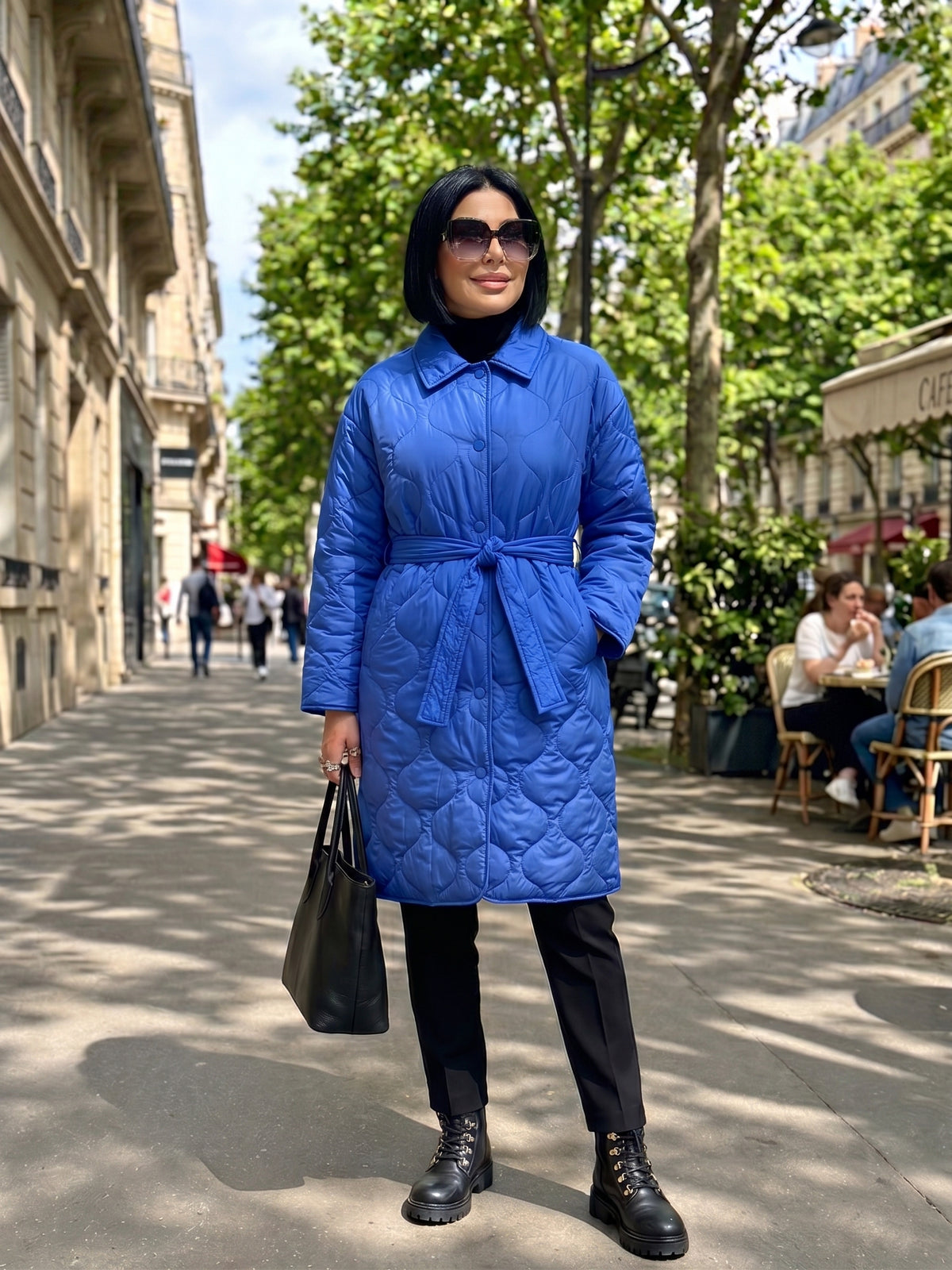 A woman stands on a sunny city sidewalk wearing a bright royal blue quilted coat with a tie waist, black pants, black boots, and holding a black tote bag.