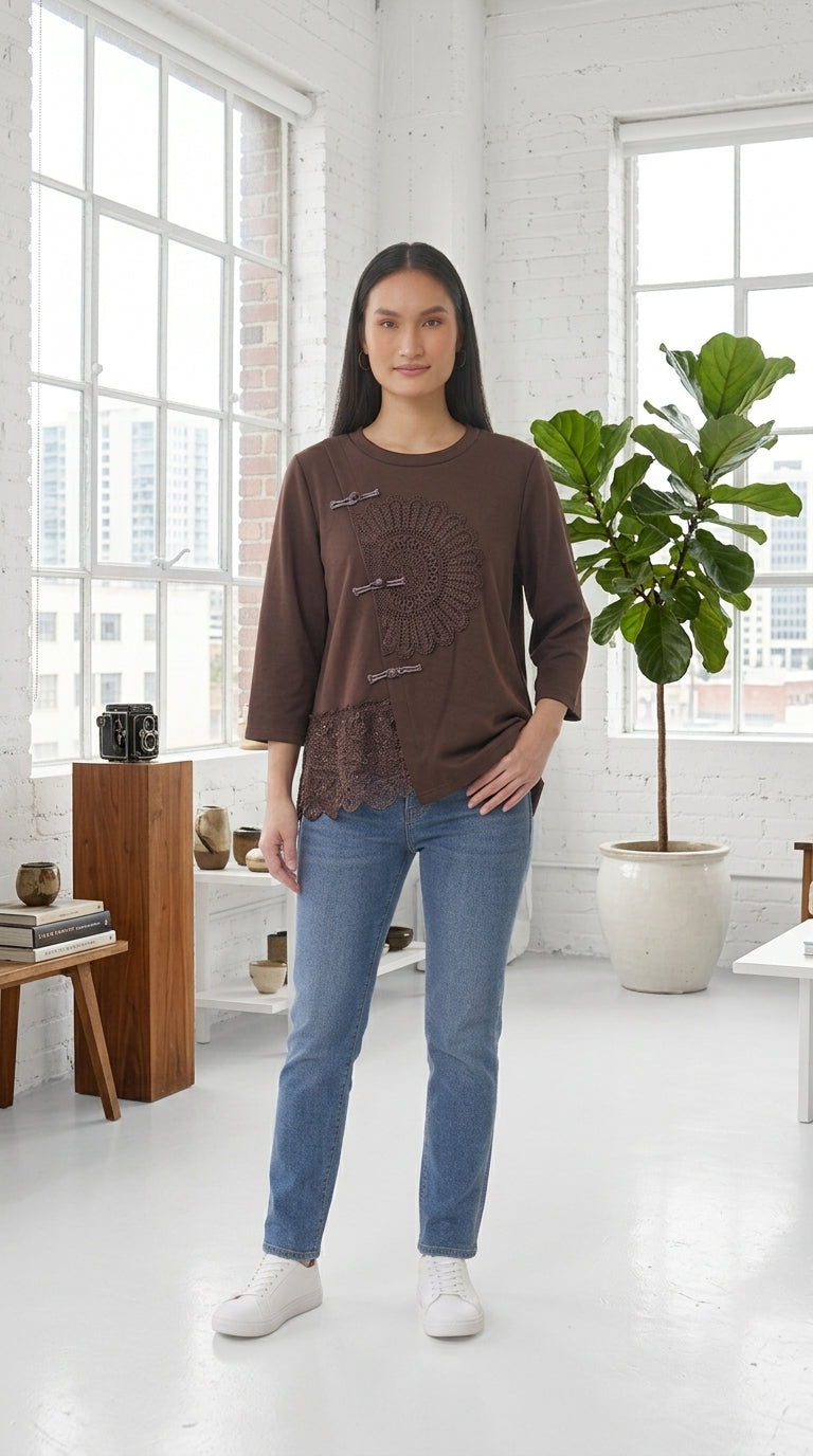 A woman modeling a brown long-sleeve t-shirt with a circular graphic print on the front, paired with blue jeans and white sneakers, standing in a bright, modern room with large windows and a potted plant.