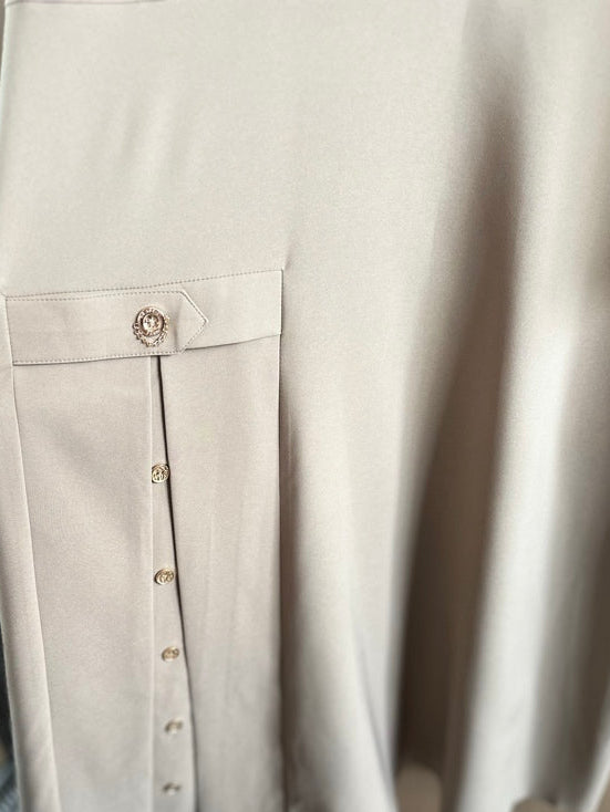 A close-up of a beige skirt with a buttoned front pocket detail, held by a hand with a paperclip. The skirt features a vertical row of gold-tone buttons and a matching waistband button.